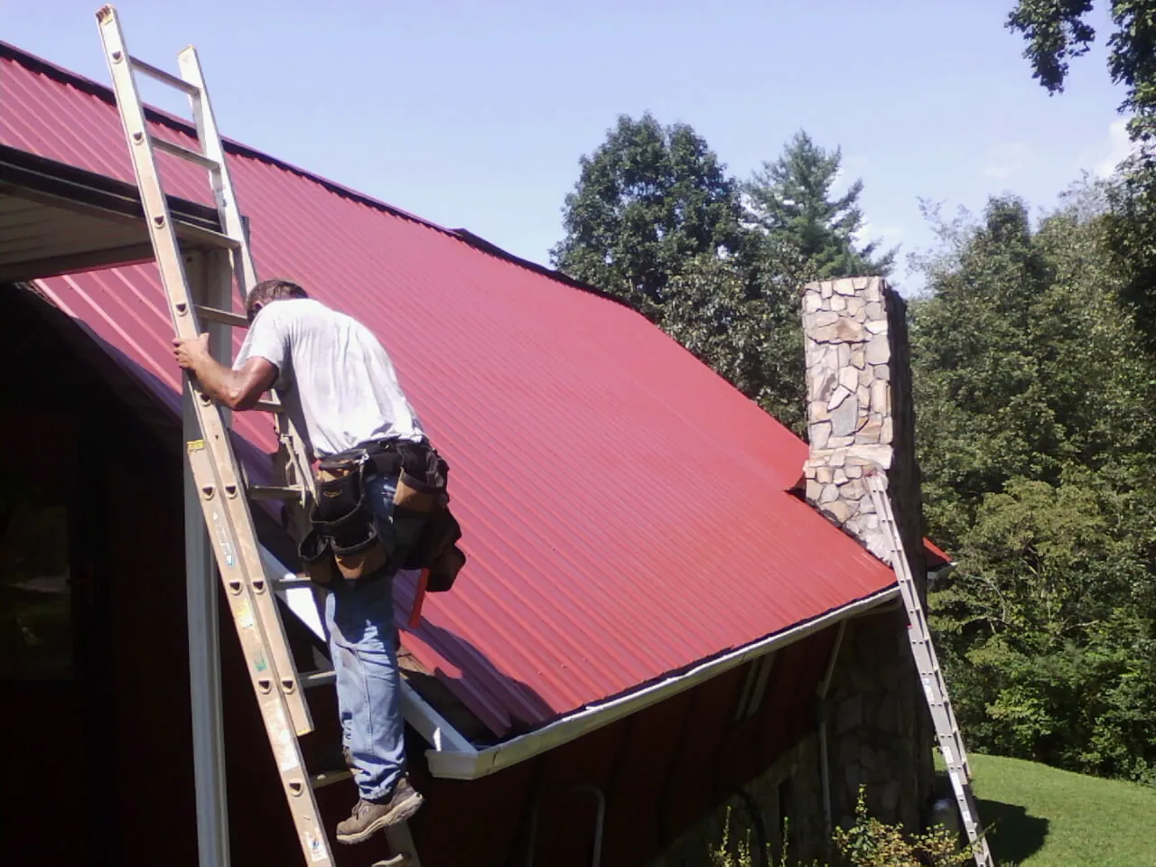 Roofer on a ladder beside a red metal roof and stone chimney — Soffit & Fascia Repair by Roofing Brothers in Ansonville